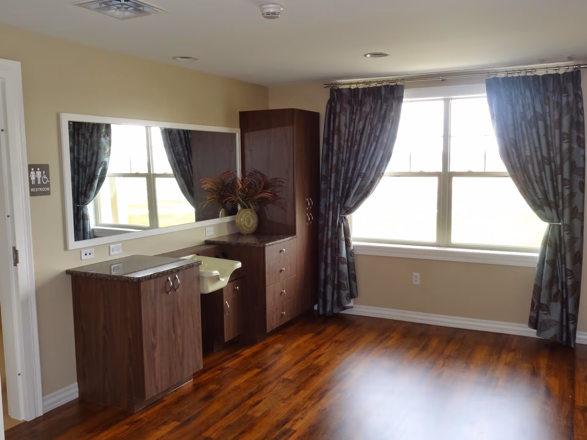 An interior room with wooden flooring, beige walls, and a large window with blue patterned curtains. There is a long mirror on the wall above a set of dark wood cabinets and a small sink. A restroom sign is visible on the left side near a door.