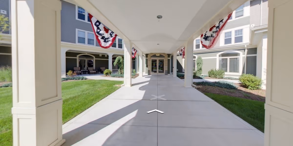 Covered walkway leading to the entrance of a residential building with patriotic bunting decorations hanging from the ceiling. The building has multiple windows and landscaped greenery on both sides of the walkway.