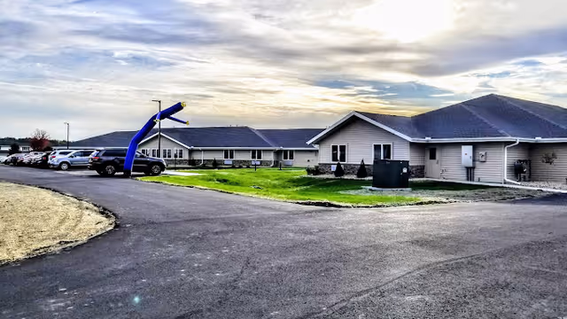 Exterior view of a single-story senior living facility building with a parking lot on the left side and a blue inflatable tube man near the entrance. The sky is partly cloudy with the sun setting or rising, casting a soft light over the scene.