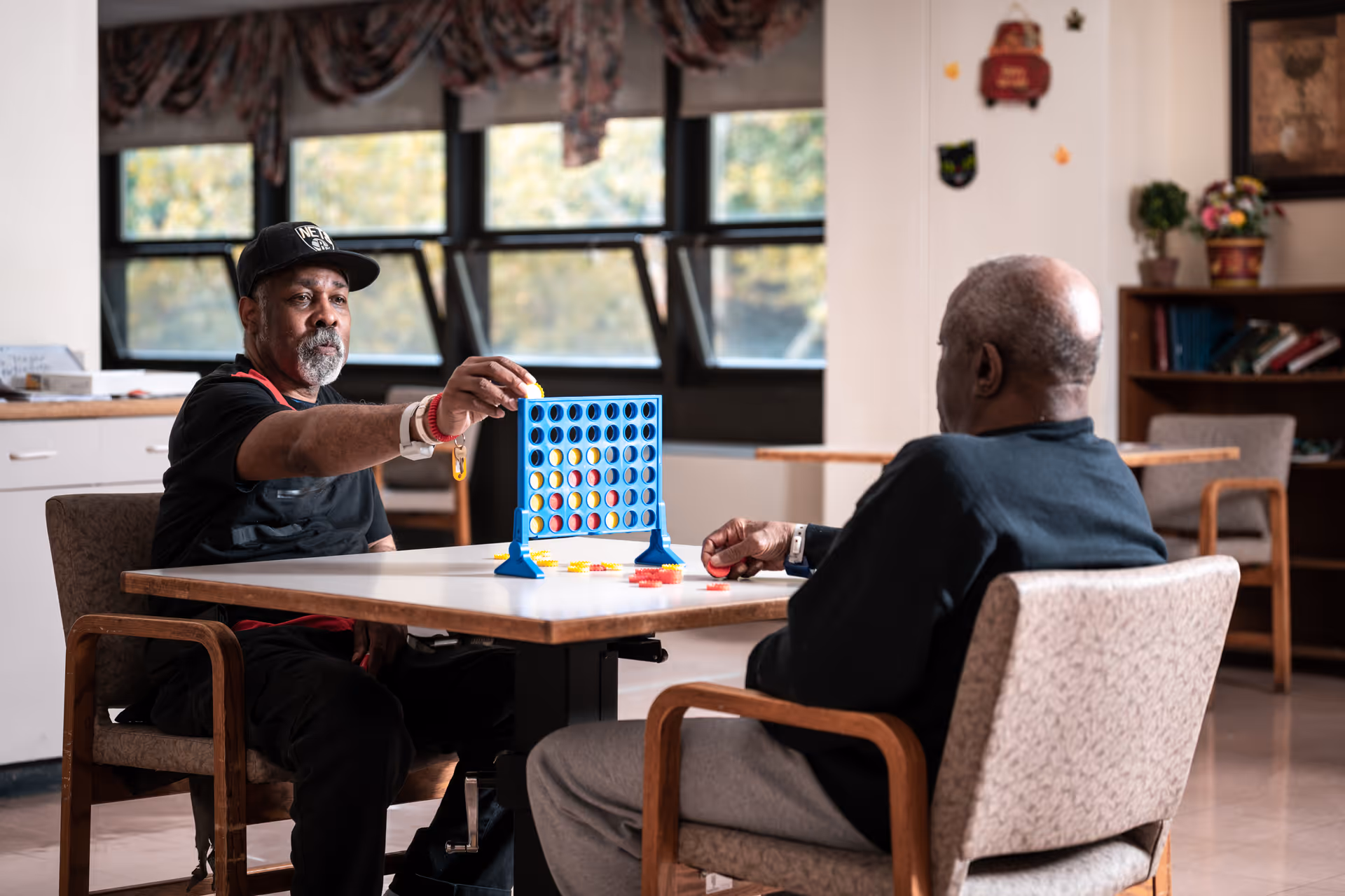 Two elderly men sitting at a table in a well-lit room playing a game of Connect Four. One man is placing a yellow game piece into the blue game grid while the other watches. The room has large windows with curtains, a bookshelf, and some wall decorations.