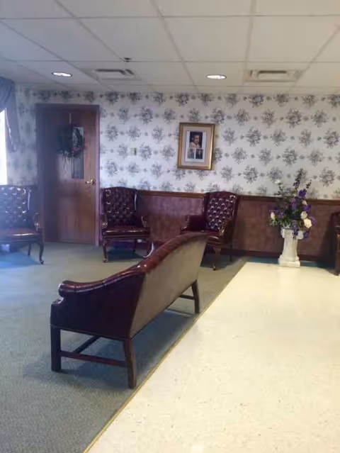 A waiting lounge with brown leather chairs, floral wallpaper, a framed photo, and vase of flowers.