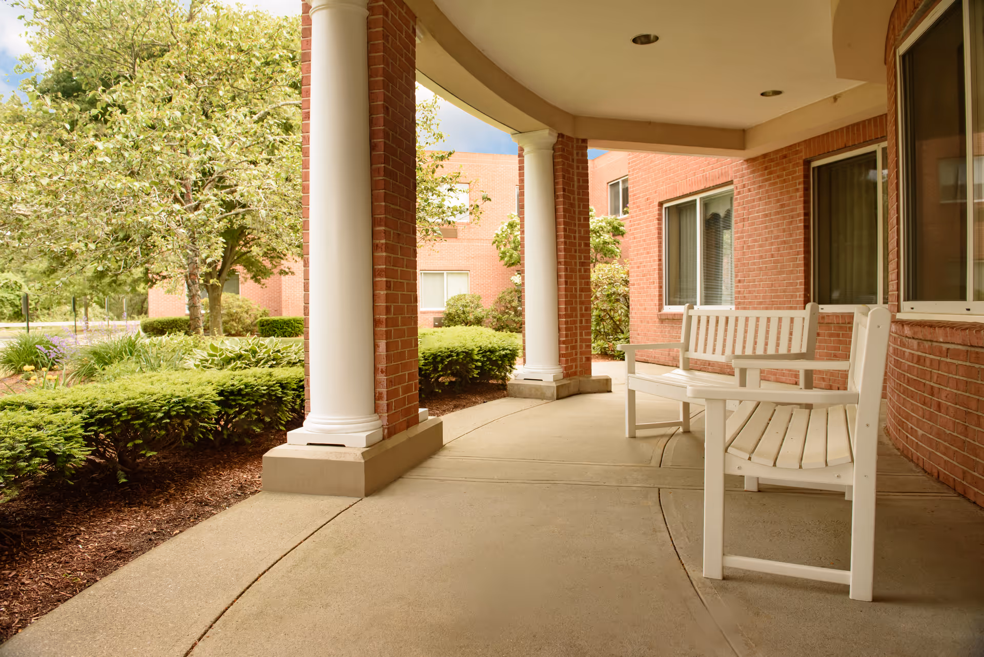 Covered outdoor patio area with white wooden benches and columns, adjacent to a red brick building with windows. Green bushes and trees surround the patio.