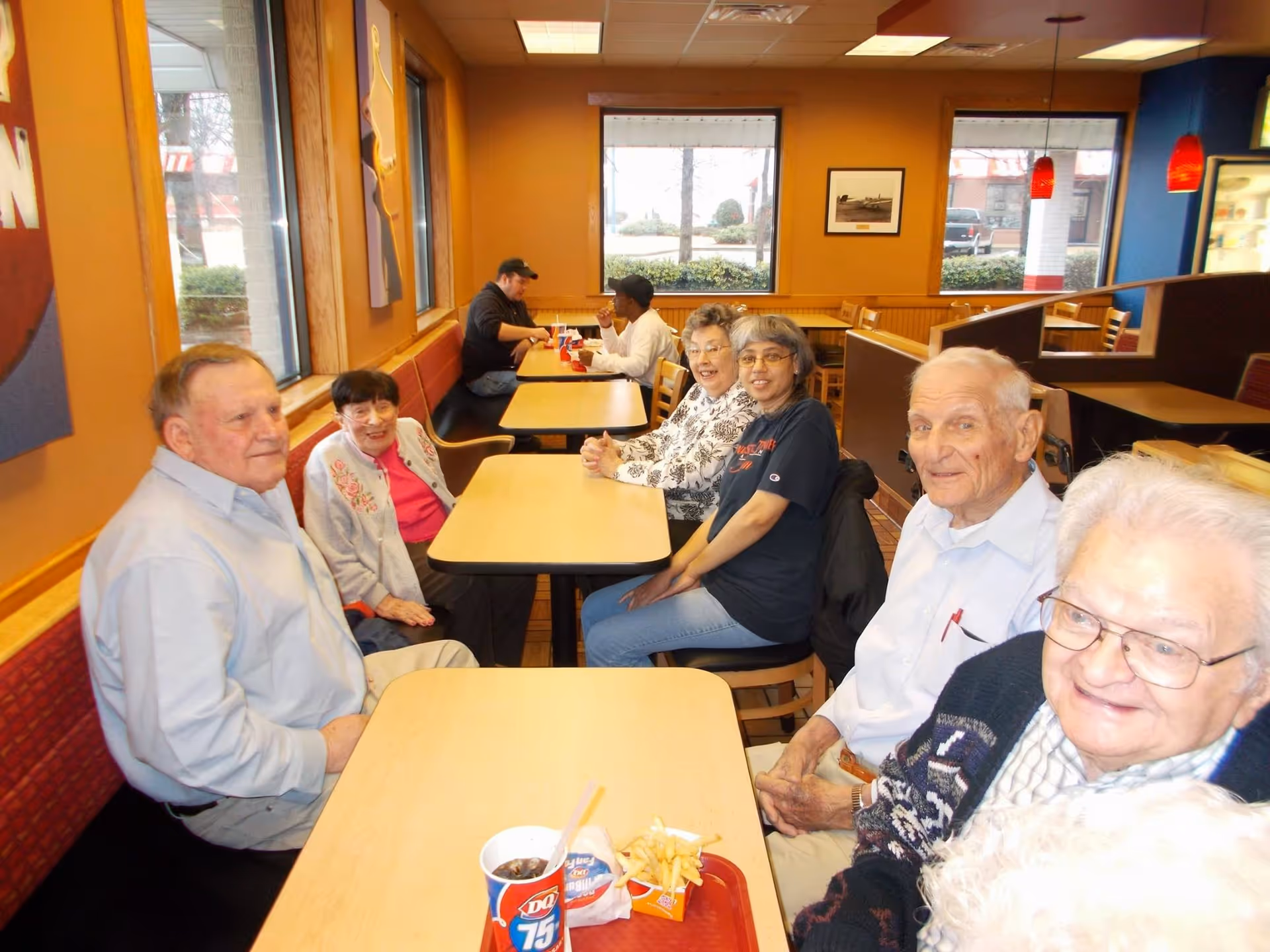 A group of elderly people sitting together at tables in a casual dining area with large windows and orange walls. Some are smiling and appear to be enjoying each other's company. There are trays with food and drinks on the tables.