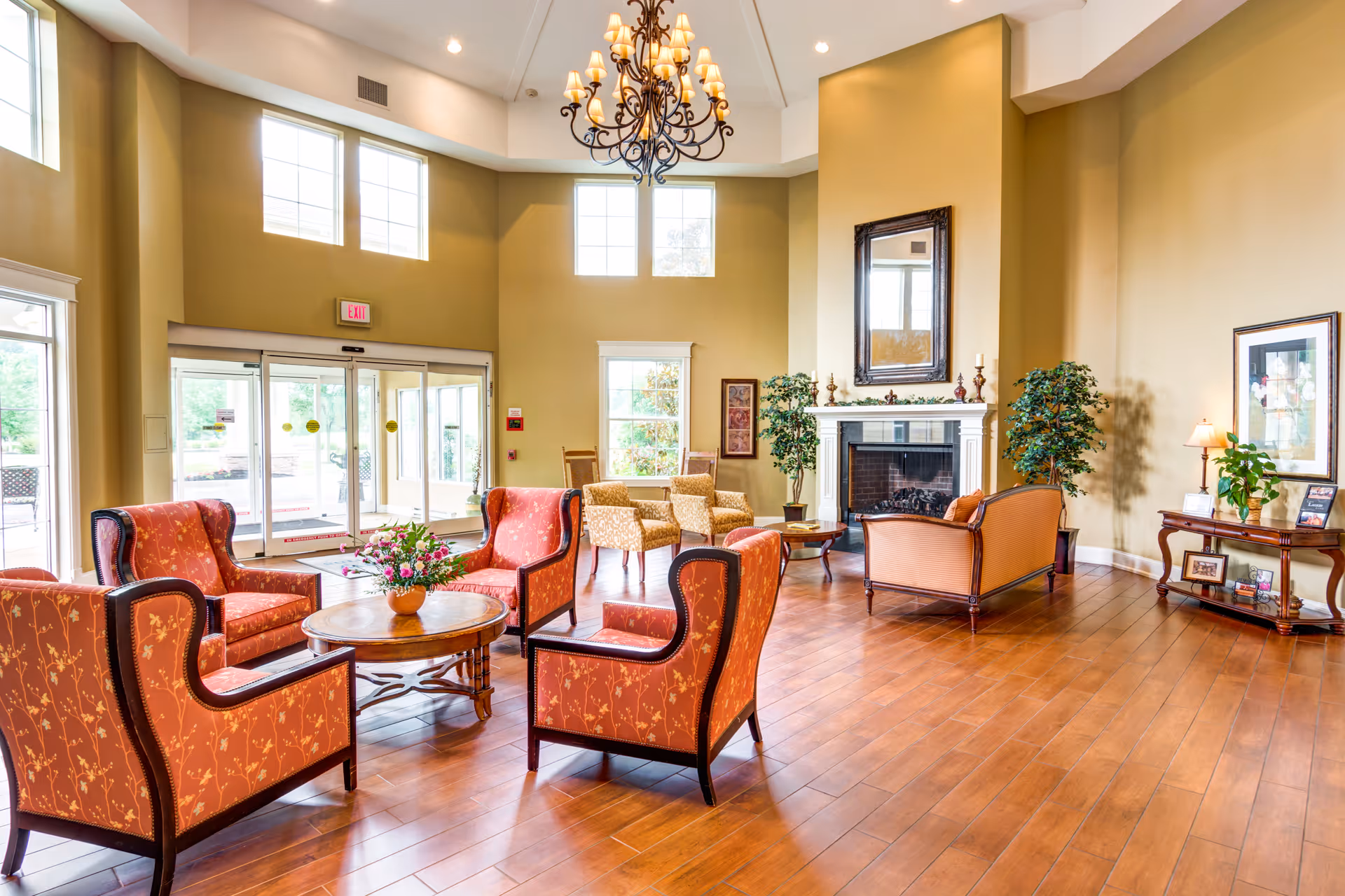 A spacious and well-lit living room area with high ceilings and large windows. The room features a chandelier, a fireplace with a mirror above it, several upholstered armchairs and sofas arranged around a wooden coffee table with a flower vase, and decorative plants. There is also a wooden side table with framed photos and a lamp. The floor is wooden, and the walls are painted a warm beige color.