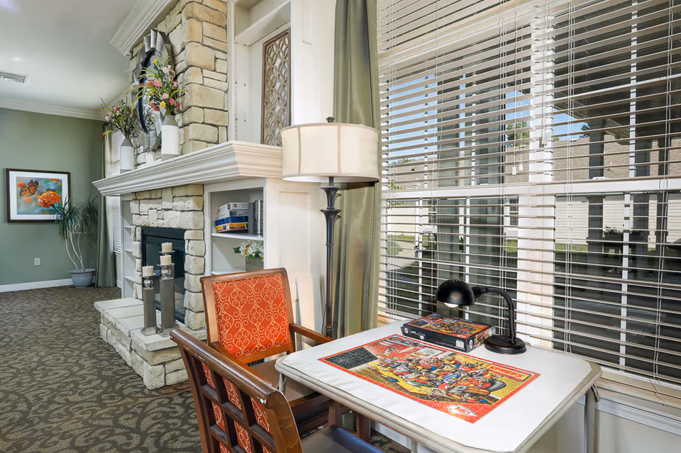 A cozy interior corner of a senior living facility featuring a stone fireplace with decorative candles and vases with flowers on the mantel. Next to the fireplace is a small table with a puzzle in progress and a box, accompanied by a wooden chair with an orange patterned cushion. A floor lamp stands beside the table, and large windows with blinds allow natural light to fill the room. The walls are painted green and decorated with a framed butterfly artwork and a potted plant.
