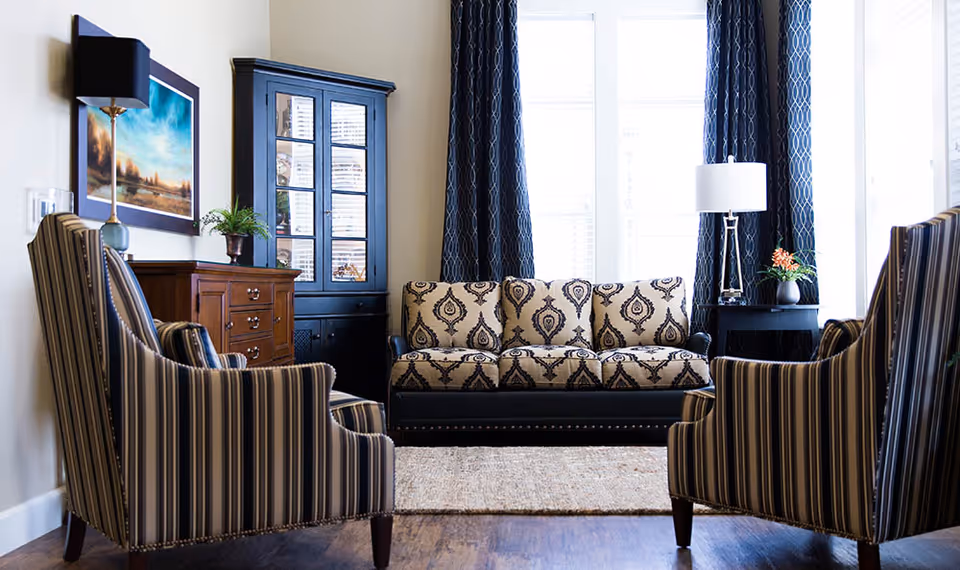 A bright living room with a patterned three-seat sofa centered between two large windows with dark curtains. Two striped armchairs face the sofa. There is a wooden cabinet with drawers and a glass-front hutch on the left side, and a small black side table with a lamp and a plant on the right side. A framed landscape painting and a wall-mounted lamp are on the left wall. The floor is wooden with a light-colored rug in front of the sofa.
