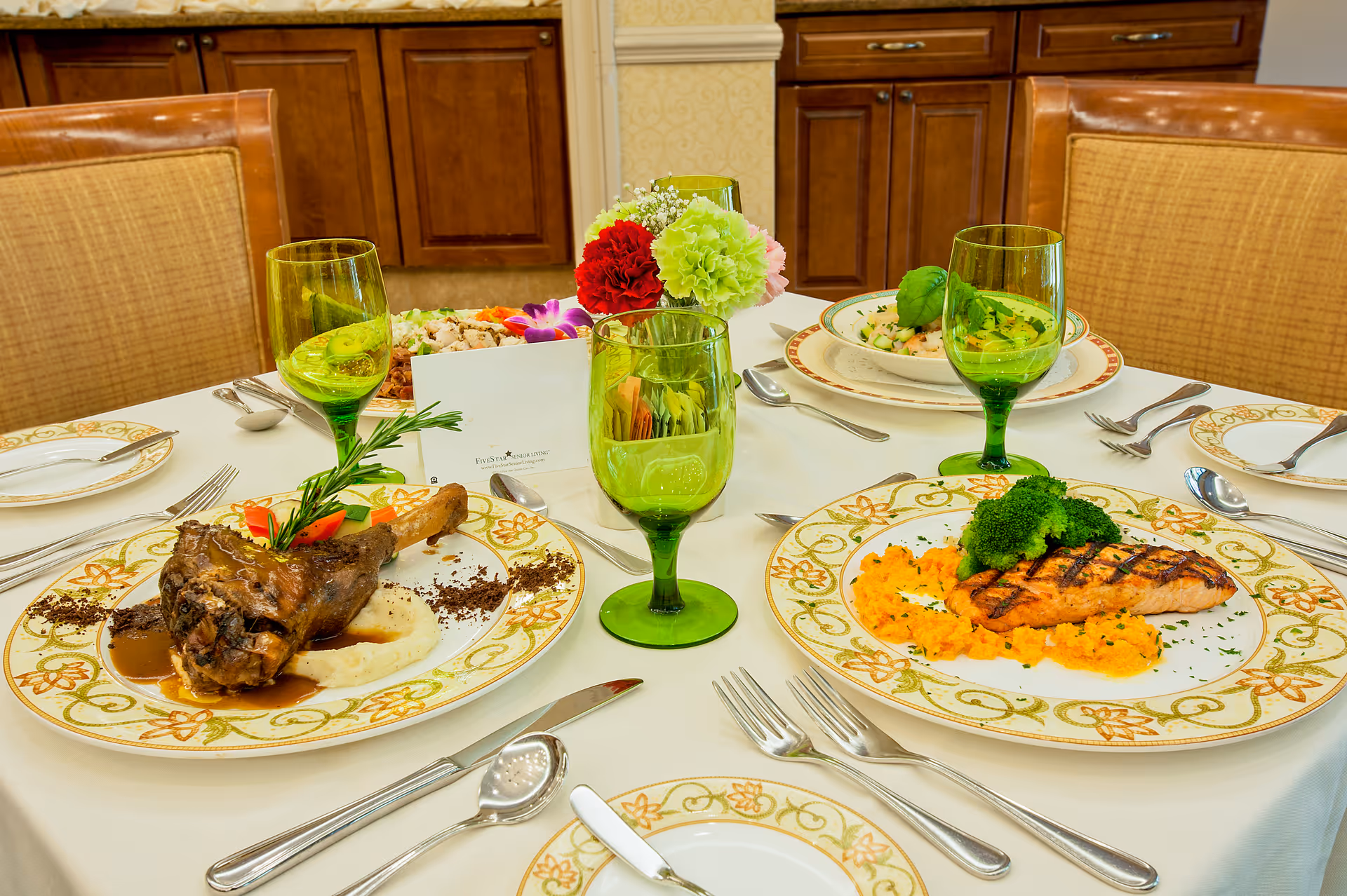 A dining table set with plated entrees, green goblets, and a floral centerpiece in a dining room.