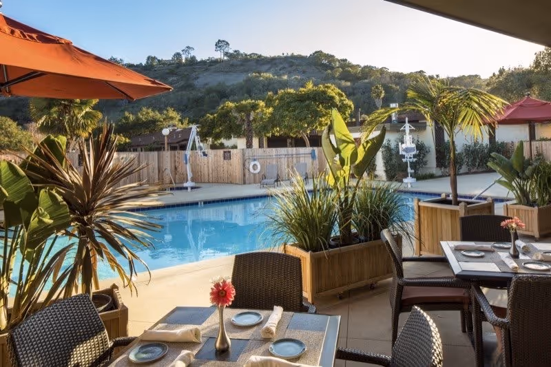 Outdoor seating area with tables set for dining near a swimming pool surrounded by plants and umbrellas, with hills and trees in the background under a clear sky.