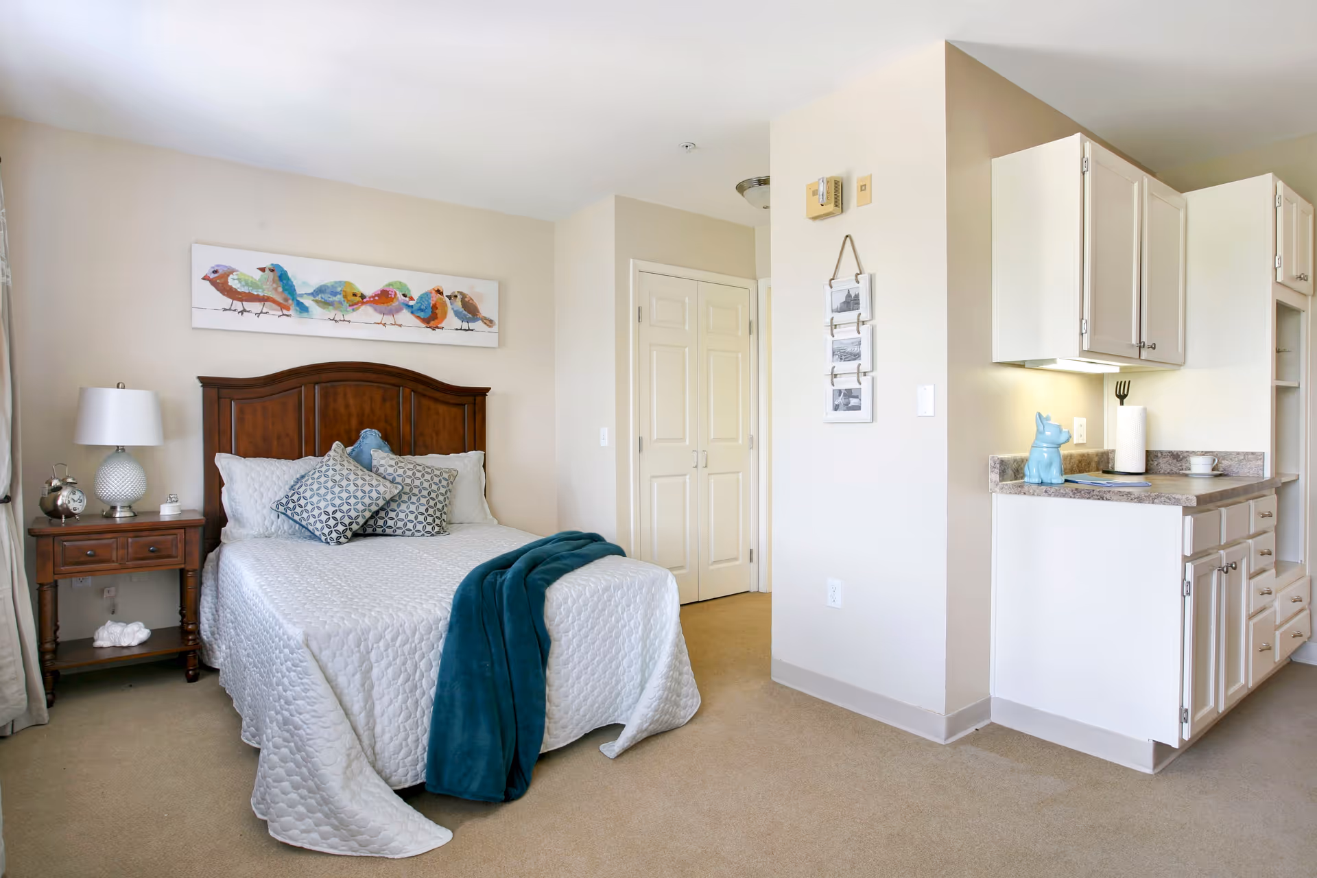 A cozy bedroom with a wooden headboard bed dressed in white quilted bedding and decorative pillows. A teal throw blanket is draped over the bed. To the left of the bed is a wooden nightstand with a white lamp and a clock. Above the bed is a colorful painting of birds. To the right, there is a small kitchenette area with white cabinets and a countertop holding a blue pig-shaped container, paper towel holder, and a cup. The room has beige walls and carpet, with a closed double-door closet in the background.