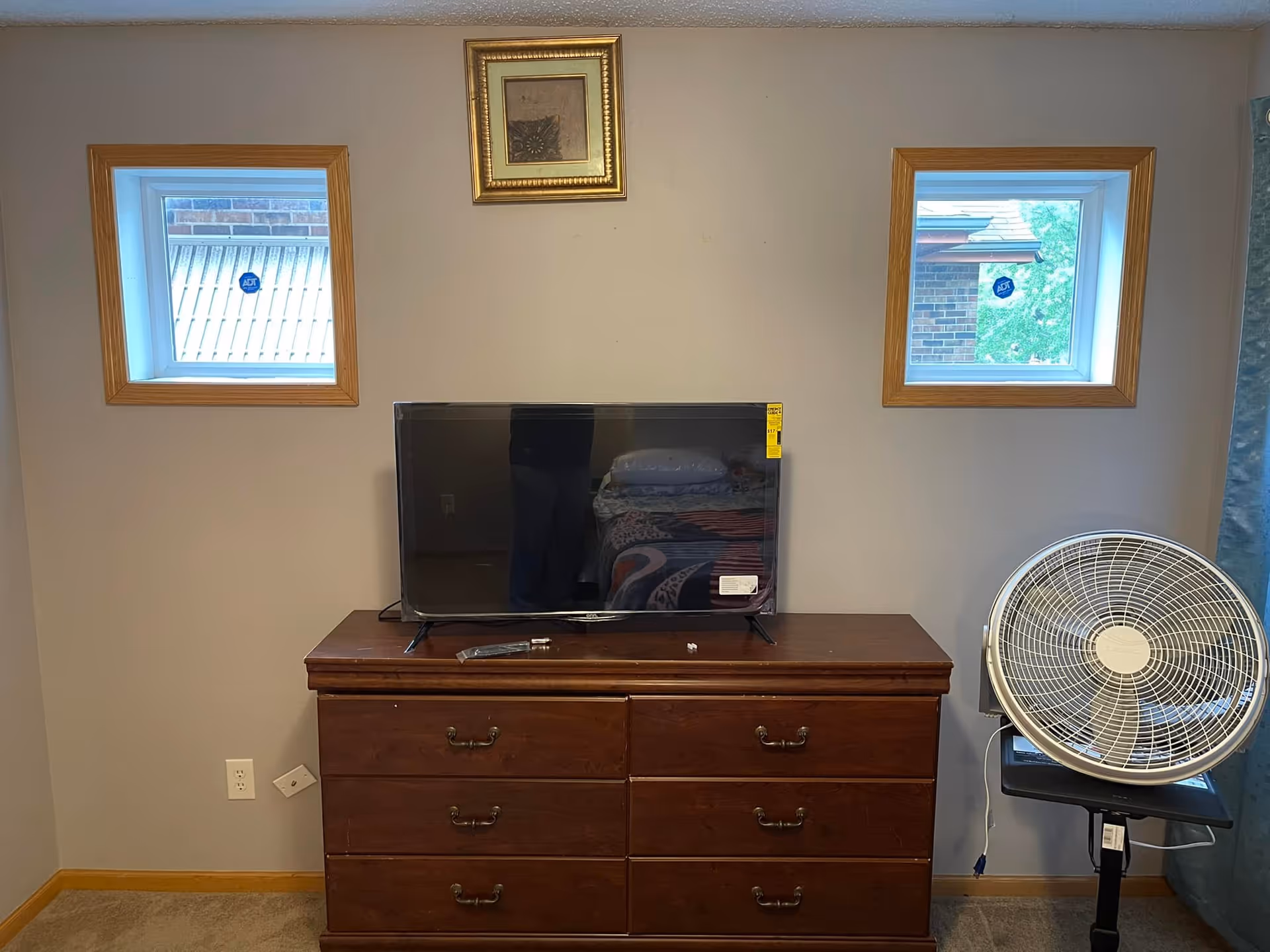 A bedroom interior showing a wooden dresser with six drawers, a flat-screen TV on top, and a standing fan to the right. There are two small square windows with wooden frames on the wall behind the dresser, and a small framed picture hanging between the windows.