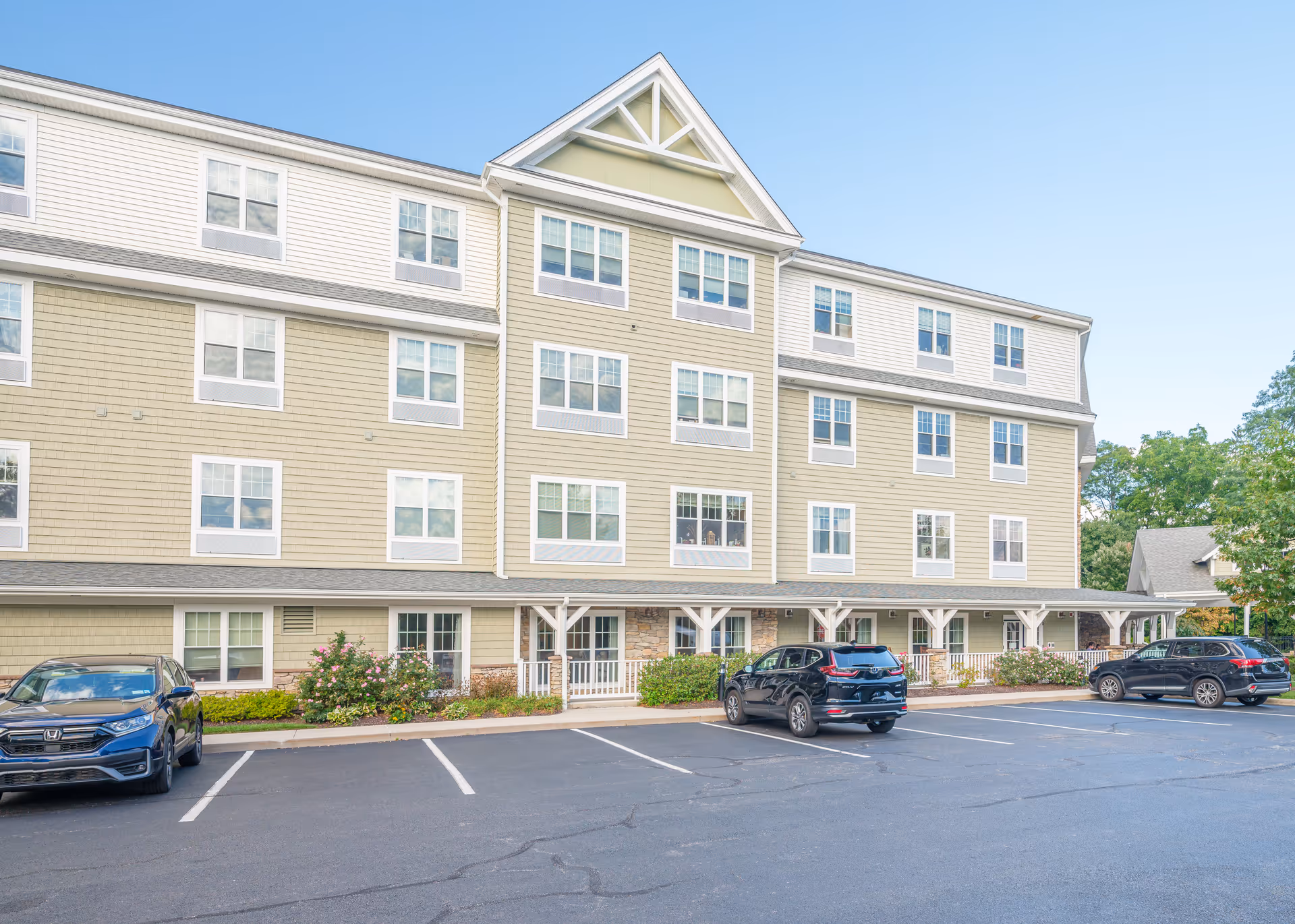 Exterior view of a multi-story senior living facility building with beige siding and white trim. There are several windows and a covered porch area at the entrance. In front of the building is a parking lot with three parked cars and some landscaping with bushes and flowers.