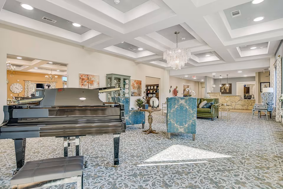 A spacious and elegant common area in a senior living facility featuring a black grand piano with a matching bench, two blue upholstered armchairs, a green leather sofa, a round wooden side table, and a chandelier hanging from a coffered ceiling. The room has patterned carpet flooring, light-colored walls with artwork, and large windows with curtains allowing natural light to enter.
