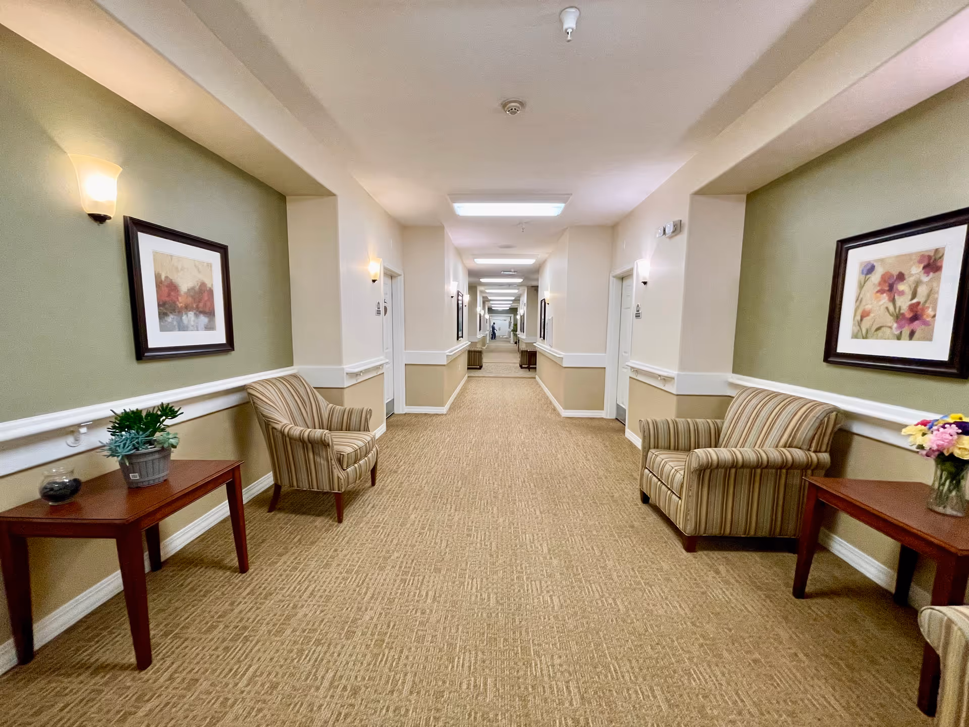 A long, well-lit hallway in an assisted living facility with beige carpet and light green and cream walls. The hallway is furnished with striped armchairs and wooden side tables holding plants and flowers. Framed floral and landscape artwork is hung on the walls, and wall sconces provide additional lighting.