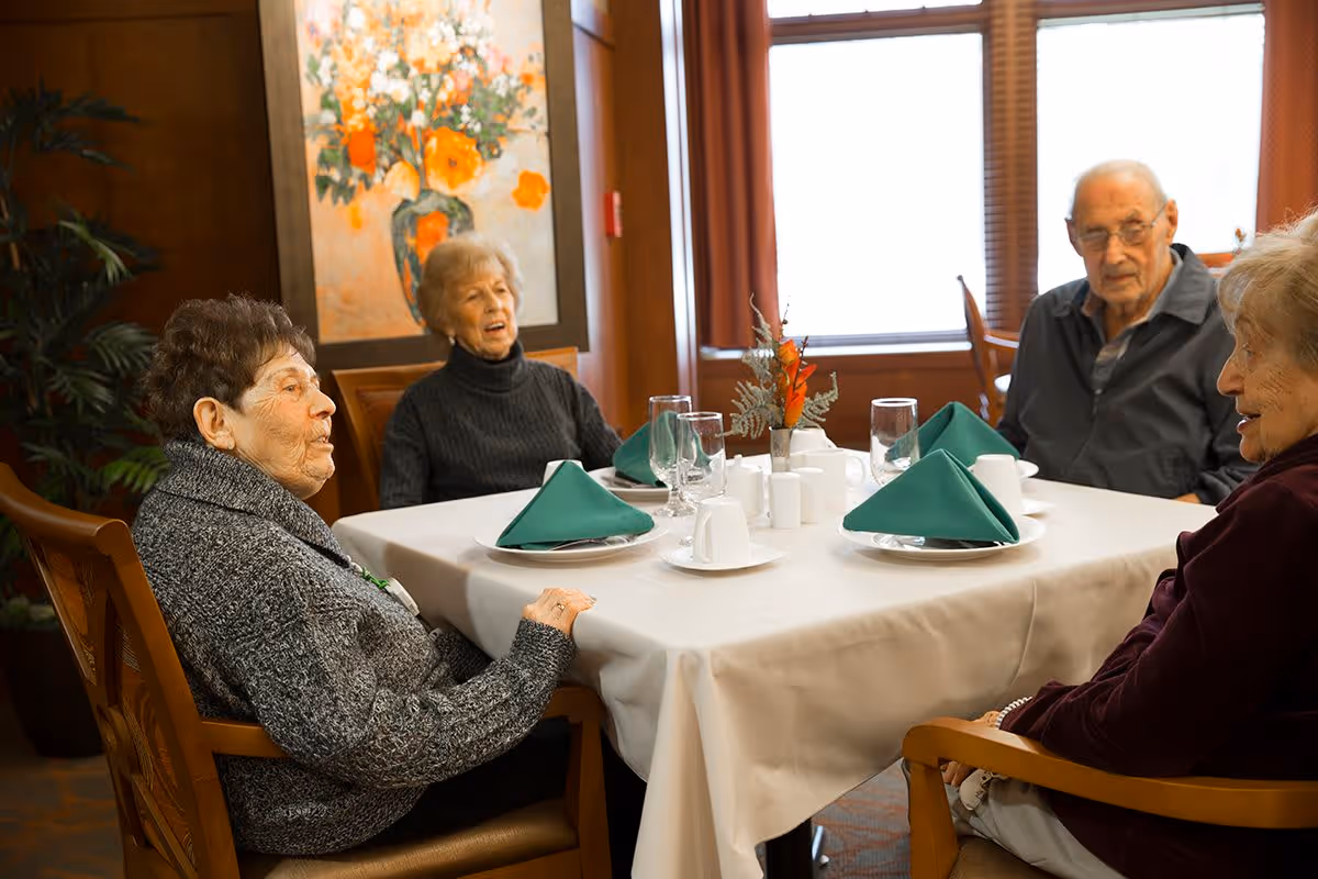 Four elderly people sitting around a dining table set with white tablecloth, green folded napkins, cups, and glasses in a well-lit room with large windows and a floral painting on the wall.