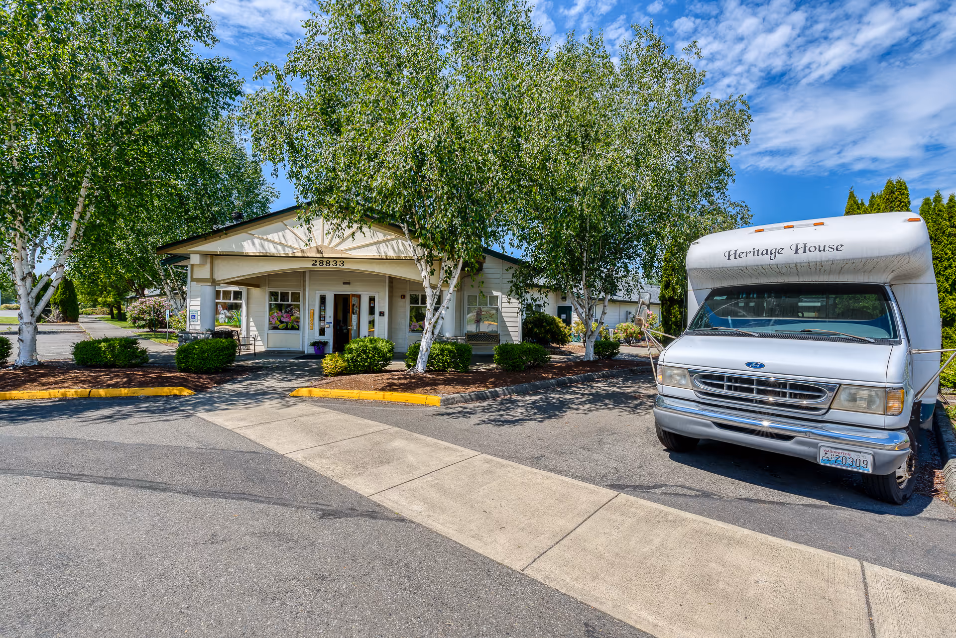 Exterior view of Heritage House Buckley Assisted Living & Memory Care building with a white shuttle bus parked in front. The building is surrounded by green trees and bushes under a blue sky with scattered clouds.