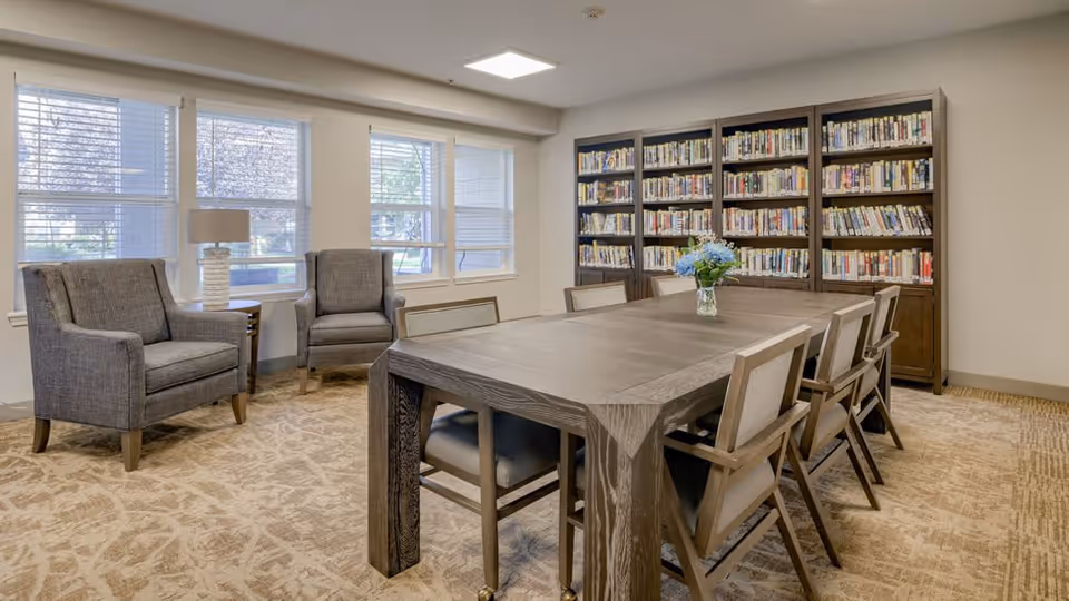 A well-lit room with a large wooden table surrounded by eight chairs. Behind the table is a tall bookshelf filled with books. To the left, there are two gray upholstered armchairs next to a window with blinds, and a small side table with a lamp. The room has beige patterned carpet and white walls.