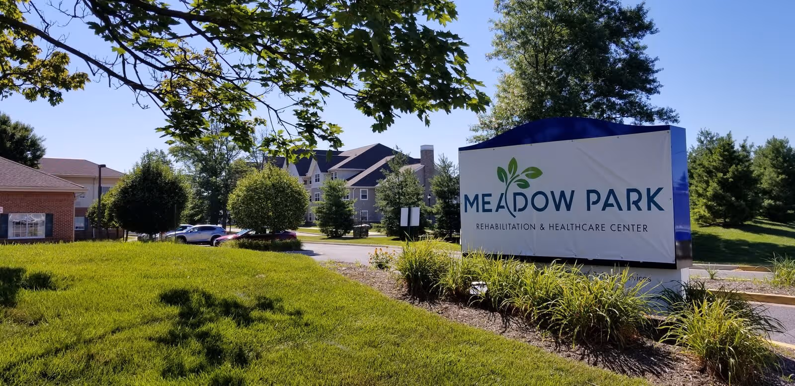Outdoor view of Meadow Park Rehabilitation and Healthcare Center sign surrounded by green grass, bushes, and trees with a clear blue sky in the background. Part of the building and parked cars are visible in the distance.