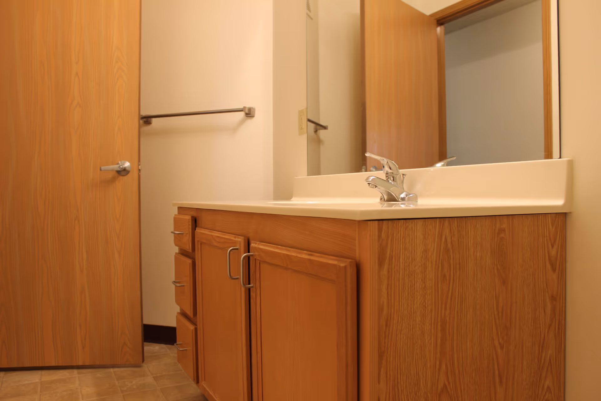 A bathroom vanity with a wooden cabinet and a white countertop with a single faucet. A large mirror is mounted above the sink, and a wooden door is partially open to the left. A towel bar is mounted on the wall next to the door.