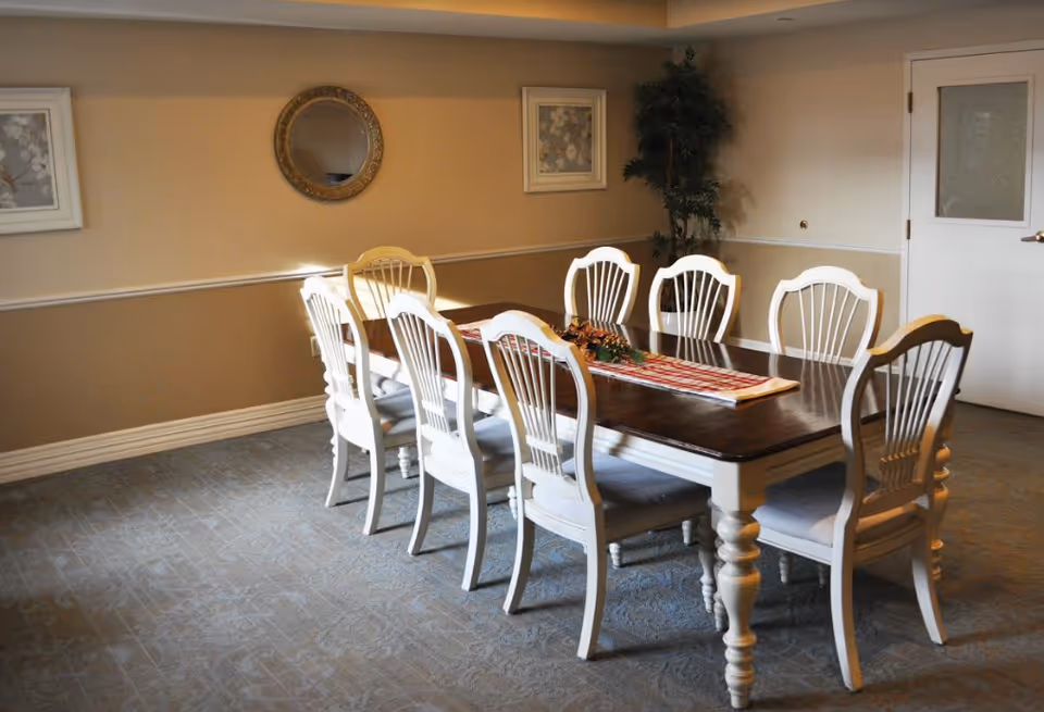 A dining room with a long wooden table and eight white chairs arranged around it. The table has a decorative runner and centerpiece. The room has beige walls with two framed pictures and a round mirror hanging on the wall. There is a potted plant in the corner and a white door with a frosted glass panel.