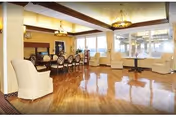 Bright communal dining and lounge area with polished wood floors, chandeliers, a long dining table and upholstered chairs facing large windows.