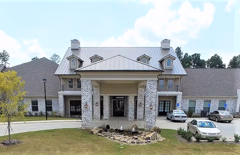 Front exterior view of The Claiborne at Hattiesburg Assisted Living building with a covered entrance supported by brick columns, a small landscaped area with rocks and a water feature in front, and several parked cars in the driveway.