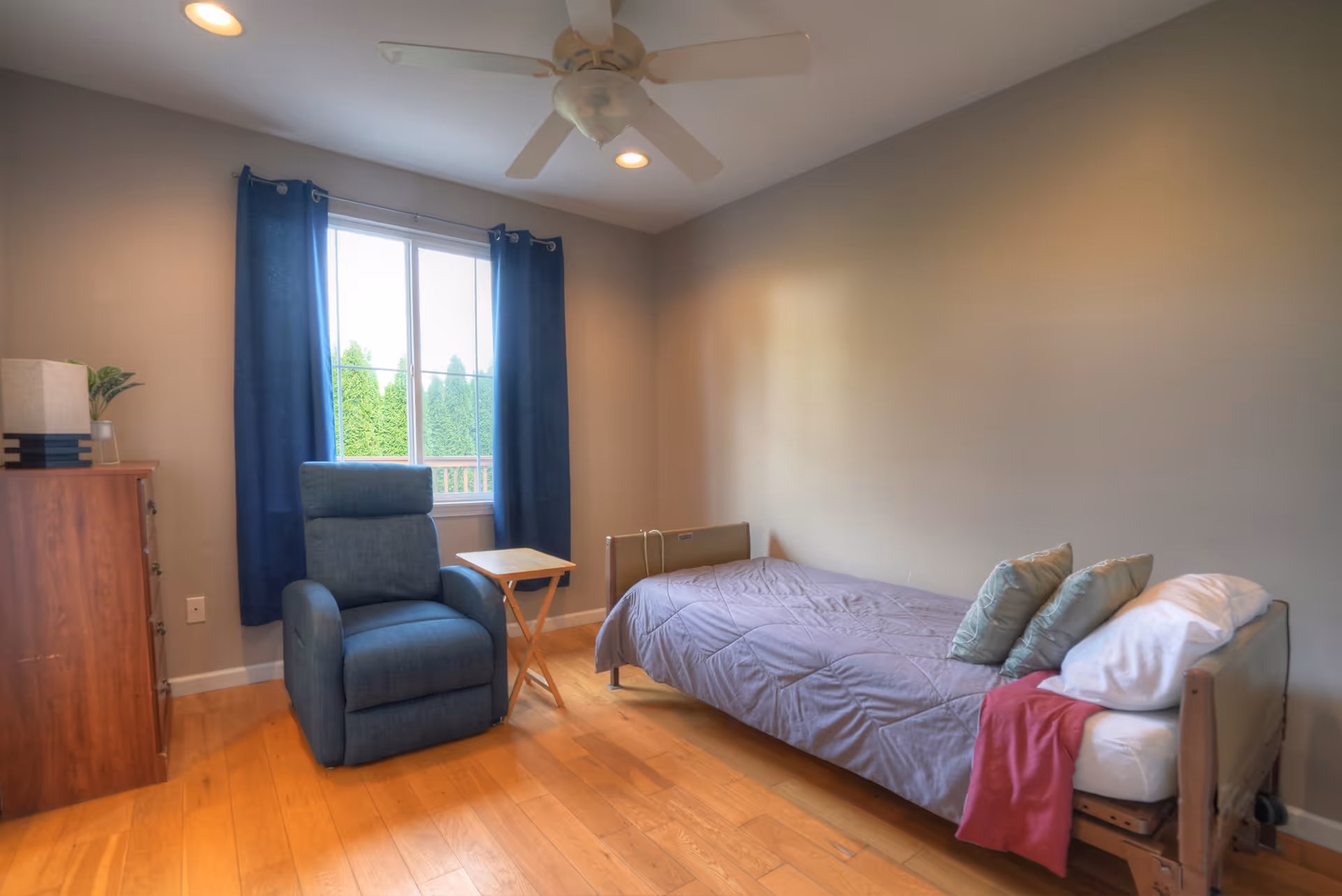 A simple bedroom with a single bed covered in a gray blanket and several pillows, a blue recliner chair, a small wooden side table, a wooden dresser with a lamp and a small plant, and a window with blue curtains letting in natural light. The room has light-colored walls, wooden flooring, and a ceiling fan with lights.