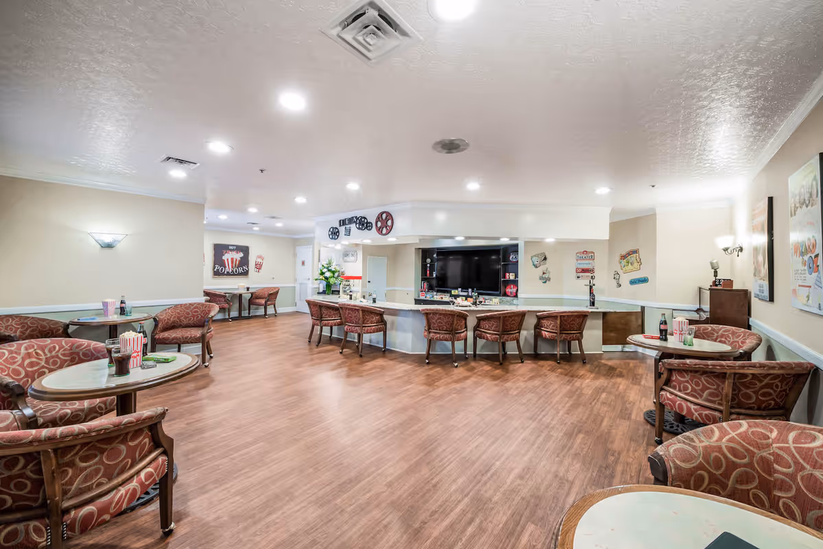 A spacious common area with wooden flooring and beige walls, featuring several round tables with patterned red chairs arranged around them. A long counter with more chairs faces a large flat-screen TV mounted on the wall. The room is decorated with movie-themed posters and wall art, including popcorn and film reel designs. The ceiling has recessed lighting and air vents.