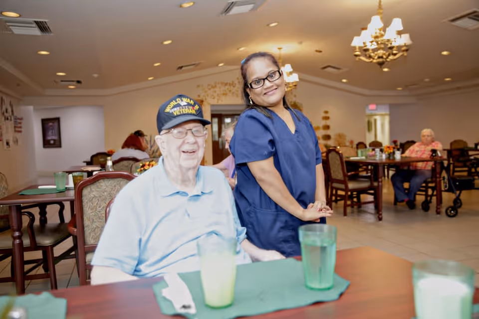An elderly man wearing a World War II veteran cap sits at a dining table with a green placemat and glass of water. A smiling female caregiver in blue scrubs stands beside him. In the background, other elderly residents sit at tables in a well-lit dining room with chandeliers and decorative wall art.