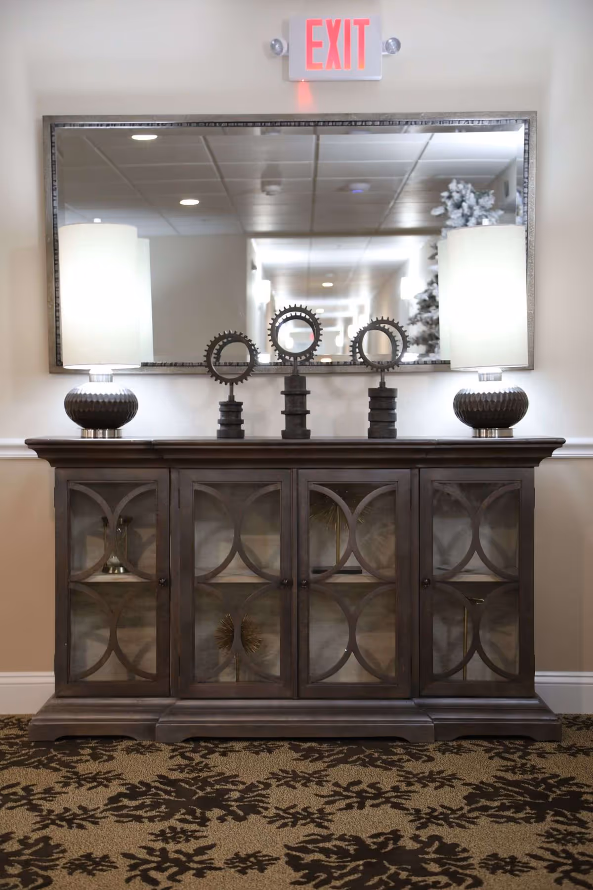 A decorative wooden cabinet with glass-paneled doors stands against a beige wall. On top of the cabinet are two matching table lamps with white shades and three circular metal gear-like sculptures. Above the cabinet is a large rectangular mirror reflecting a hallway with ceiling lights and a Christmas tree. An illuminated red EXIT sign is mounted on the wall above the mirror. The floor is carpeted with a patterned design in dark brown and beige.