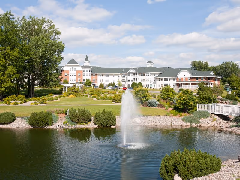 A large retirement community building with white and red brick exterior, green roofs, and multiple windows, situated behind a landscaped garden with bushes, trees, and a pond featuring a water fountain in the foreground under a partly cloudy sky.