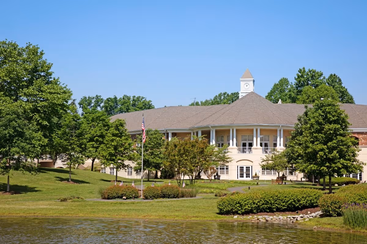 Exterior view of The Fairfax at Belvoir Woods building with a clock tower on the roof, surrounded by green trees and landscaping, with a pond in the foreground and a clear blue sky.