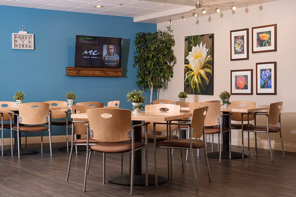 A dining area with several wooden tables and chairs arranged neatly. The walls are decorated with framed floral artwork and a large green plant is placed in the corner. A wall-mounted TV is visible on a blue accent wall, and a clock hangs nearby. Small potted plants are placed on each table.