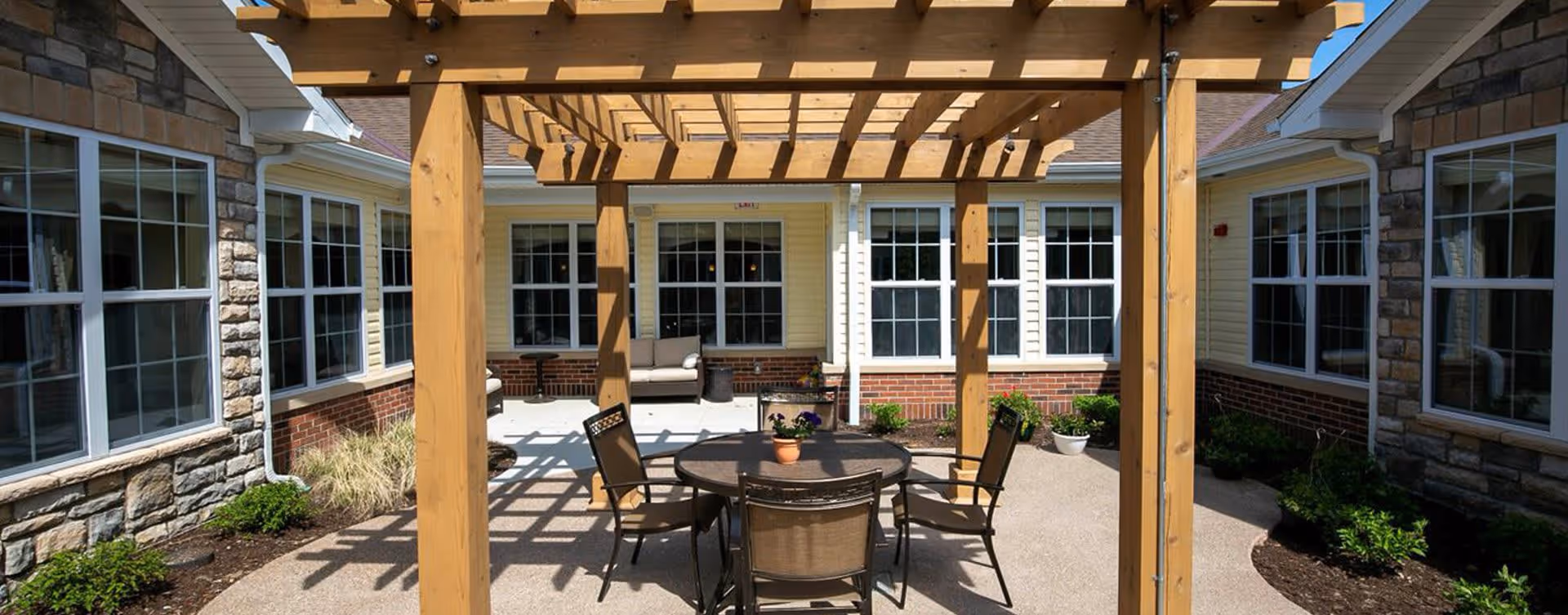 Outdoor patio area at Bickford of Midland featuring a wooden pergola with a round table and four chairs underneath. The patio is surrounded by a building with large windows, stone and brick exterior walls, and some potted plants and landscaping.