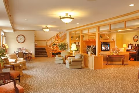 A spacious and warmly lit living room area in a senior living facility with comfortable armchairs, a patterned carpet, a staircase in the background, and a television set within a wooden partition. There are also tables with lamps and decorative plants, creating a cozy and inviting atmosphere.