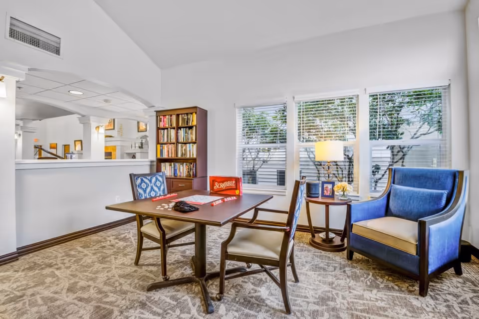A cozy sitting area with a table set up for a game of Scrabble, two chairs with patterned cushions, a blue armchair, a small round side table with a lamp and framed photo, and a bookshelf filled with books. Large windows with blinds let in natural light and show trees outside.