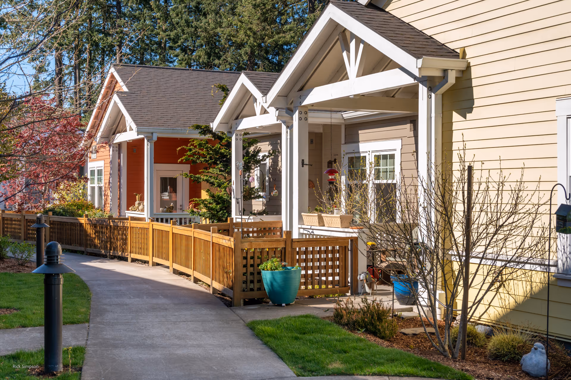 A sunny outdoor walkway lined with small residential-style buildings featuring porches, wooden fences, and landscaped greenery including trees and shrubs.