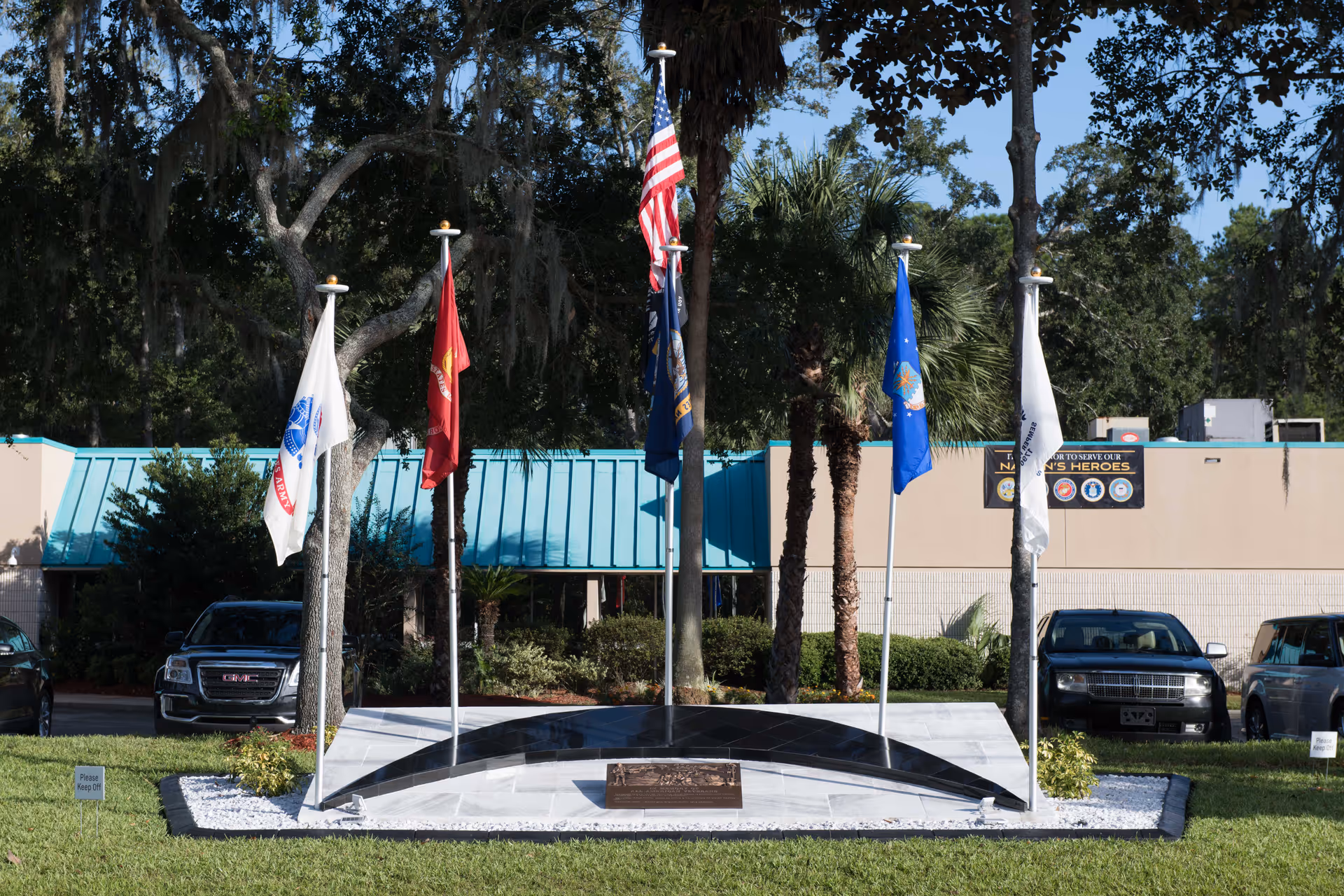 Outdoor memorial display with five flagpoles holding various flags including the American flag, set in front of a building with a teal roof and surrounded by trees and parked cars.
