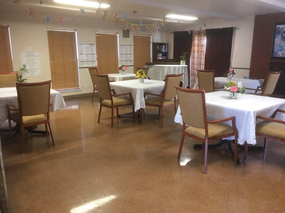Dining room with tables covered in white tablecloths, chairs, and small floral centerpieces.