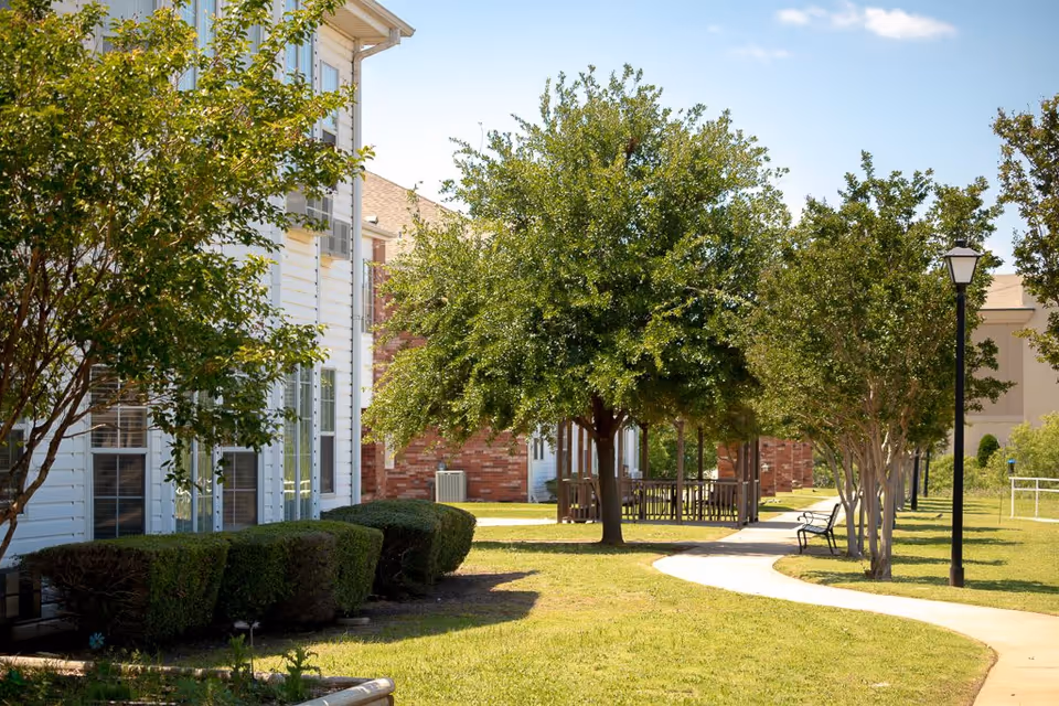 A sunny outdoor pathway lined with trees and bushes beside a white building with multiple windows. There are benches and a lamp post along the curved sidewalk, with a gazebo visible in the background.
