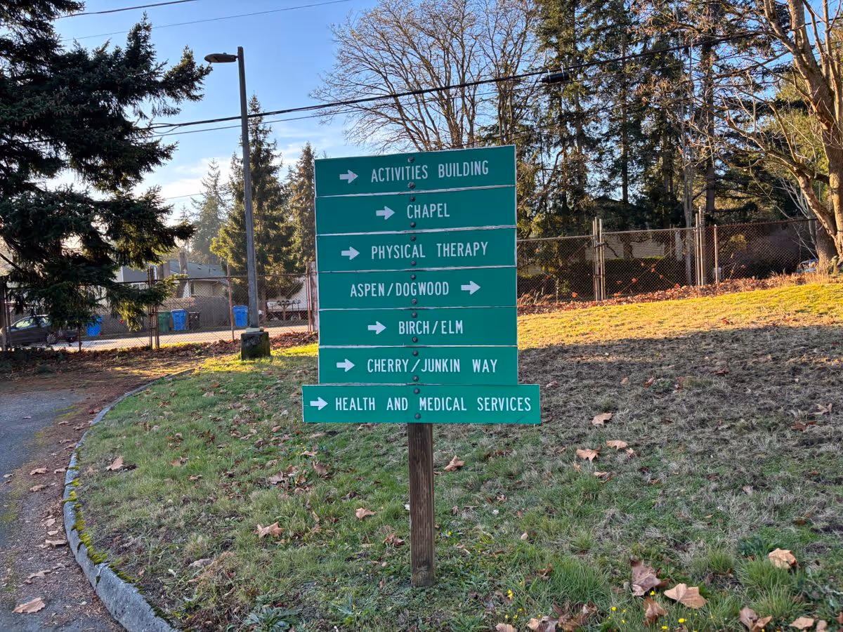 Green directional sign listing campus locations (Activities Building, Chapel, Physical Therapy, Aspen/Dogwood, Birch/Elm, Cherry/Junkin Way, Health and Medical Services) standing on a grassy area with trees and a fence in the background.