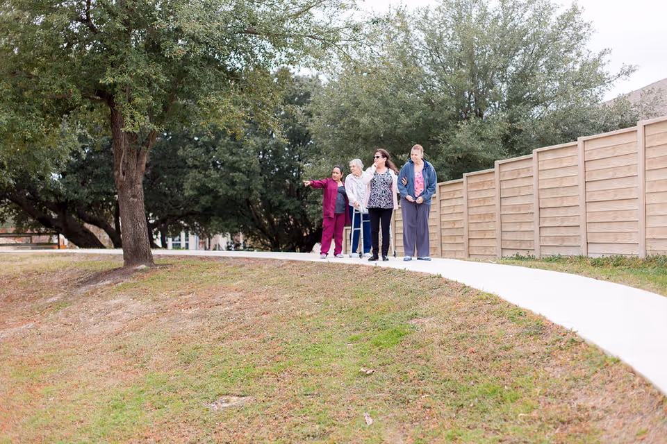 Four women walking together on a curved paved path outdoors, surrounded by grass, trees, and a wooden fence. One woman is pointing ahead while the others look in the same direction.
