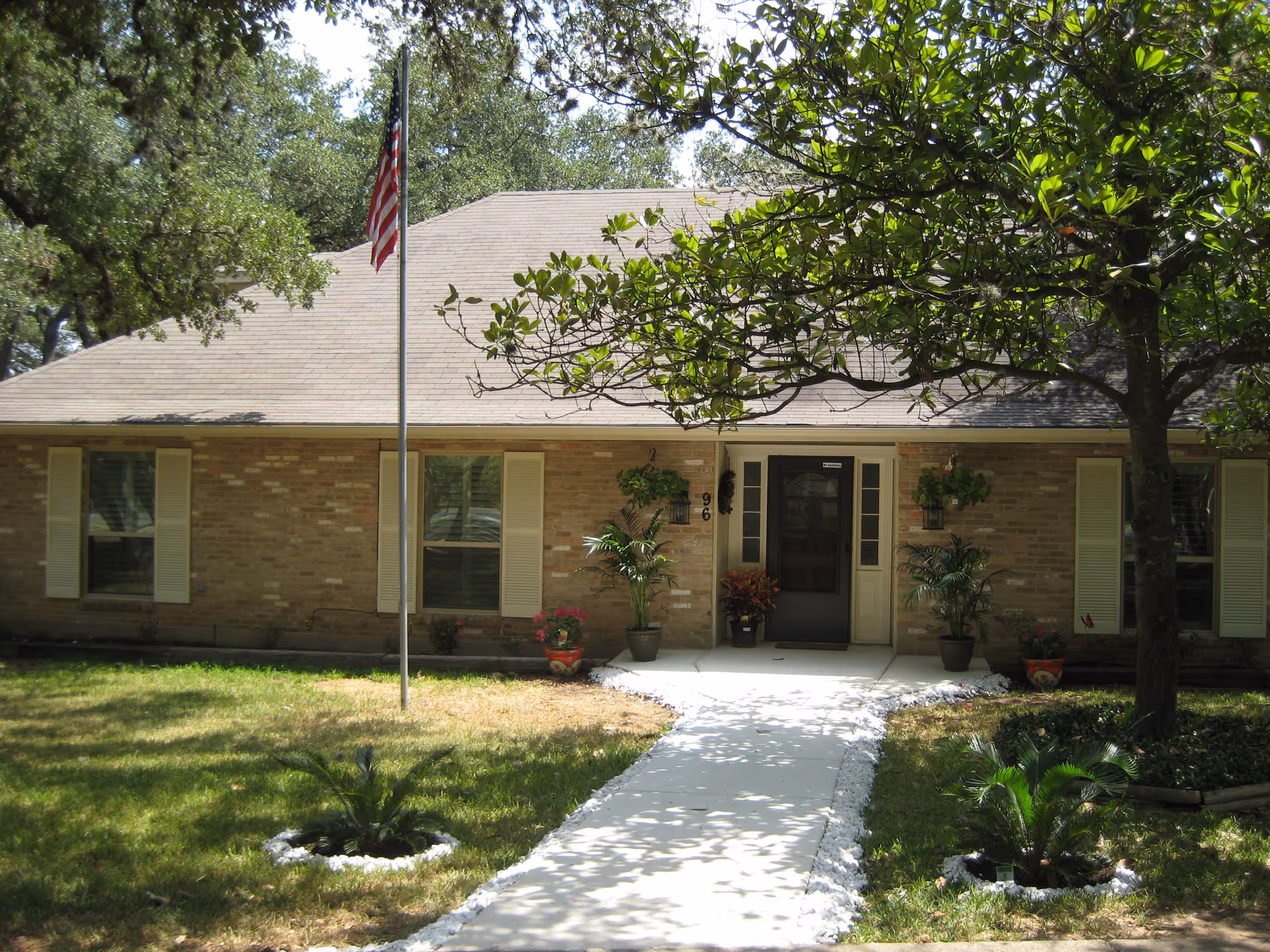 Front exterior view of a single-story brick building with a sloped roof, an American flag on a flagpole, a concrete walkway leading to a black front door, and several potted plants and trees in the yard.