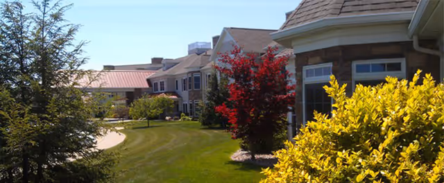 Exterior view of a senior living building with a well-kept lawn, colorful shrubs, and a paved walkway.