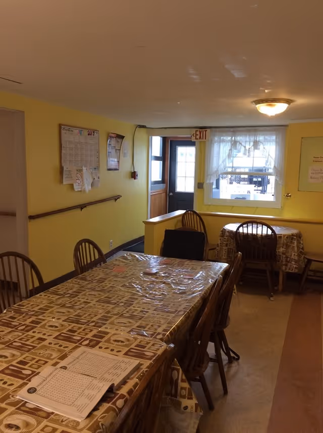 Interior view of a dining area with yellow walls, two tables covered with patterned tablecloths and plastic covers, several wooden chairs, a window with sheer curtains, an exit door, and a bulletin board on the wall.