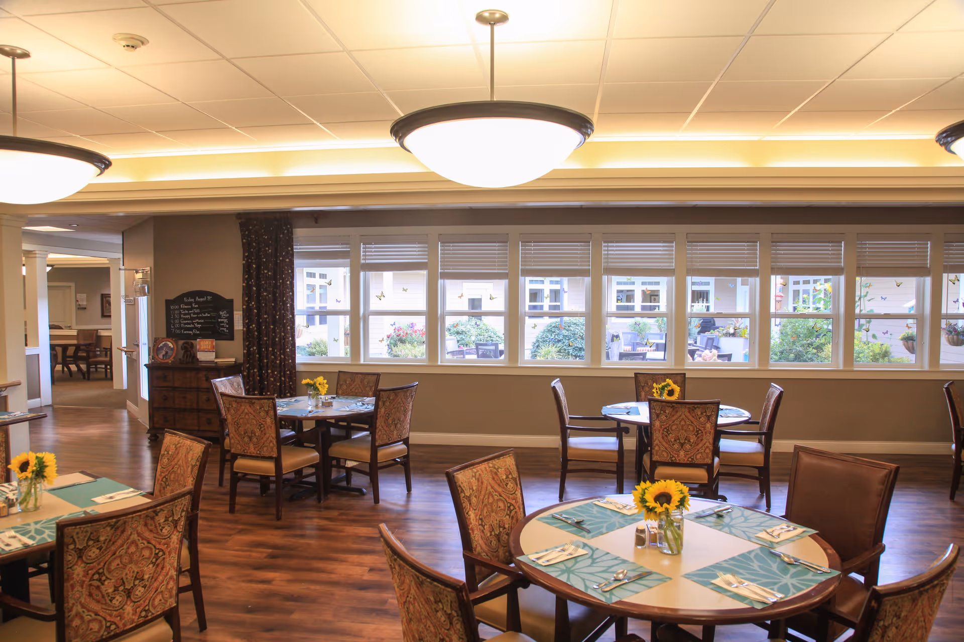 Sunlit dining room with round tables set with placemats and sunflower centerpieces and large windows overlooking a courtyard.