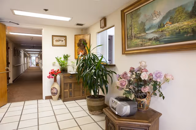 Interior hallway area of St. John Of Kronstadt Home featuring a tiled floor, a wooden cabinet with potted plants and flowers, a small stereo, and framed artwork on the walls. The hallway extends into the distance with doors and handrails along the walls.
