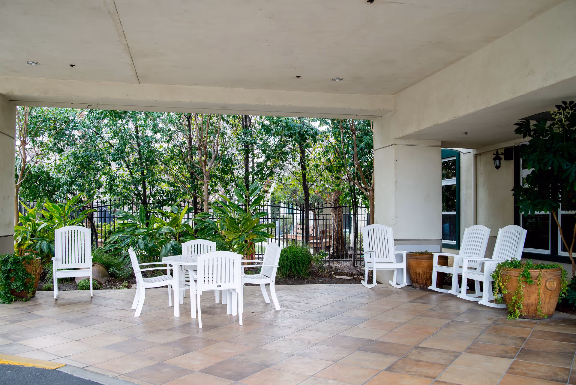 Covered outdoor patio area with white plastic chairs and a table on tiled flooring, surrounded by potted plants and greenery with a black metal fence in the background.
