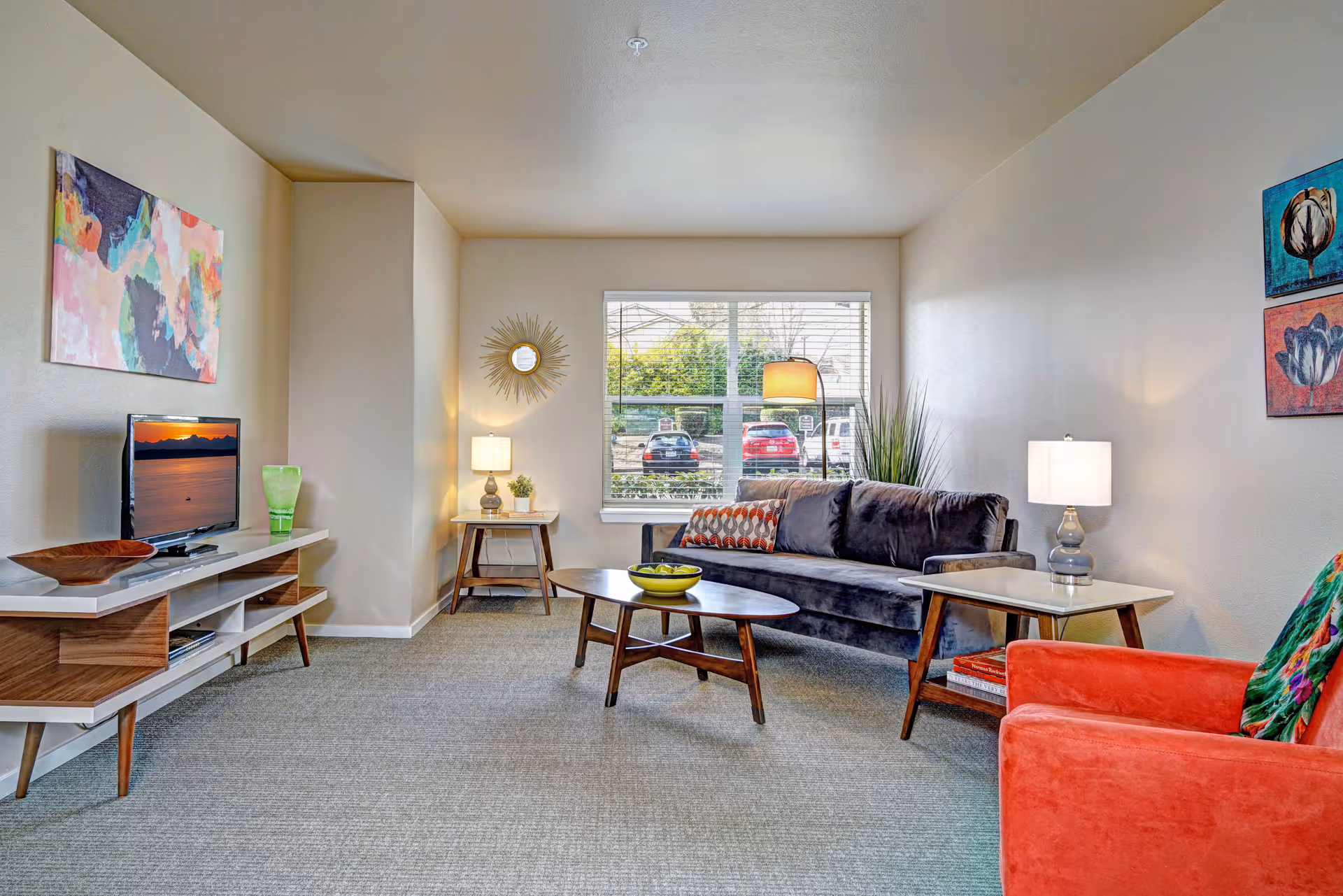 A bright and cozy living room with a gray sofa, an orange armchair, a wooden coffee table with a bowl of green apples, two side tables with lamps, a TV on a modern wooden stand, colorful wall art, and a window showing parked cars outside.