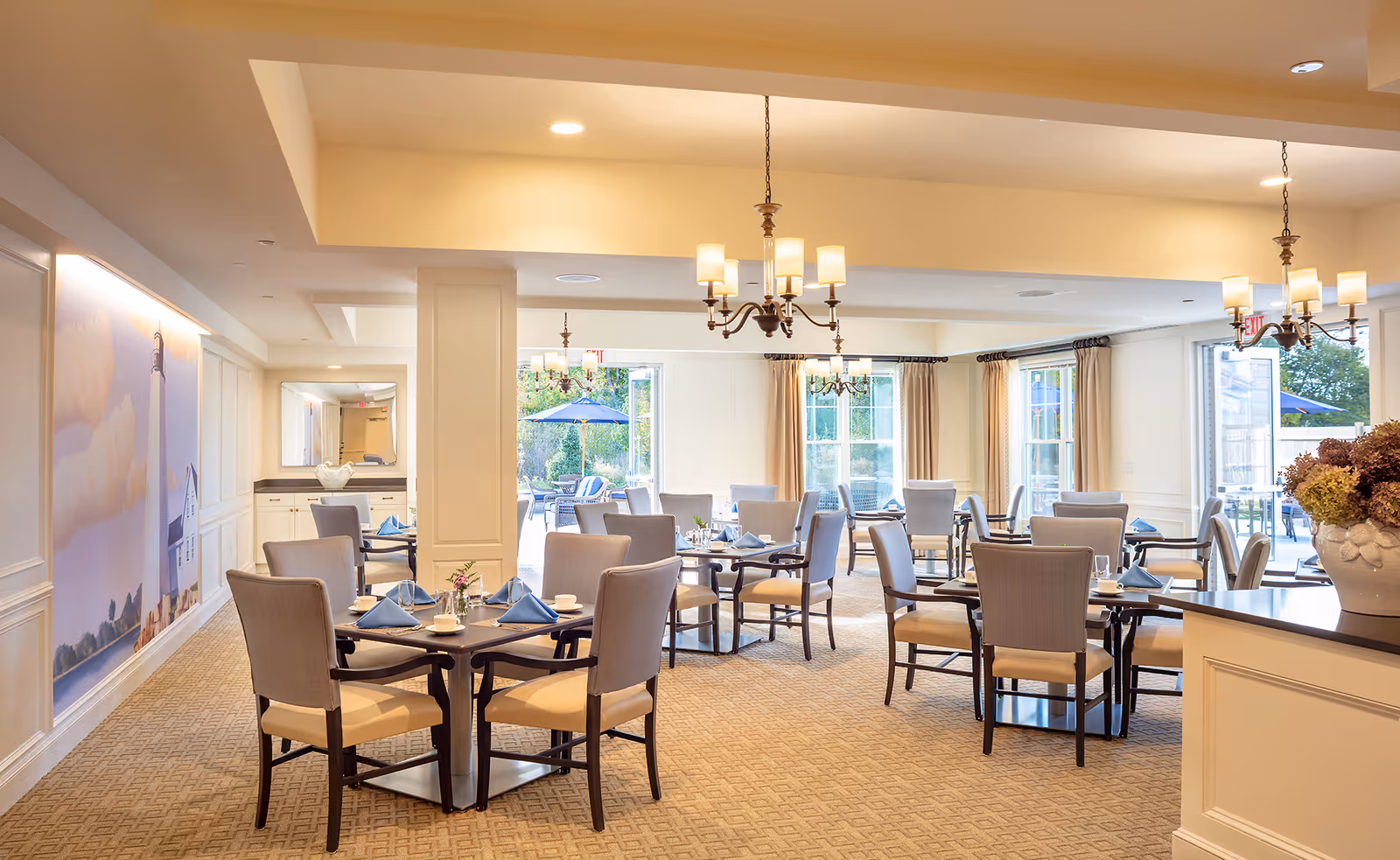 A bright and elegant dining room with multiple tables set with blue napkins, cups, and small flower arrangements. The room features beige carpeting, cream-colored walls, chandeliers, large windows with curtains, and a view of an outdoor patio with umbrellas.