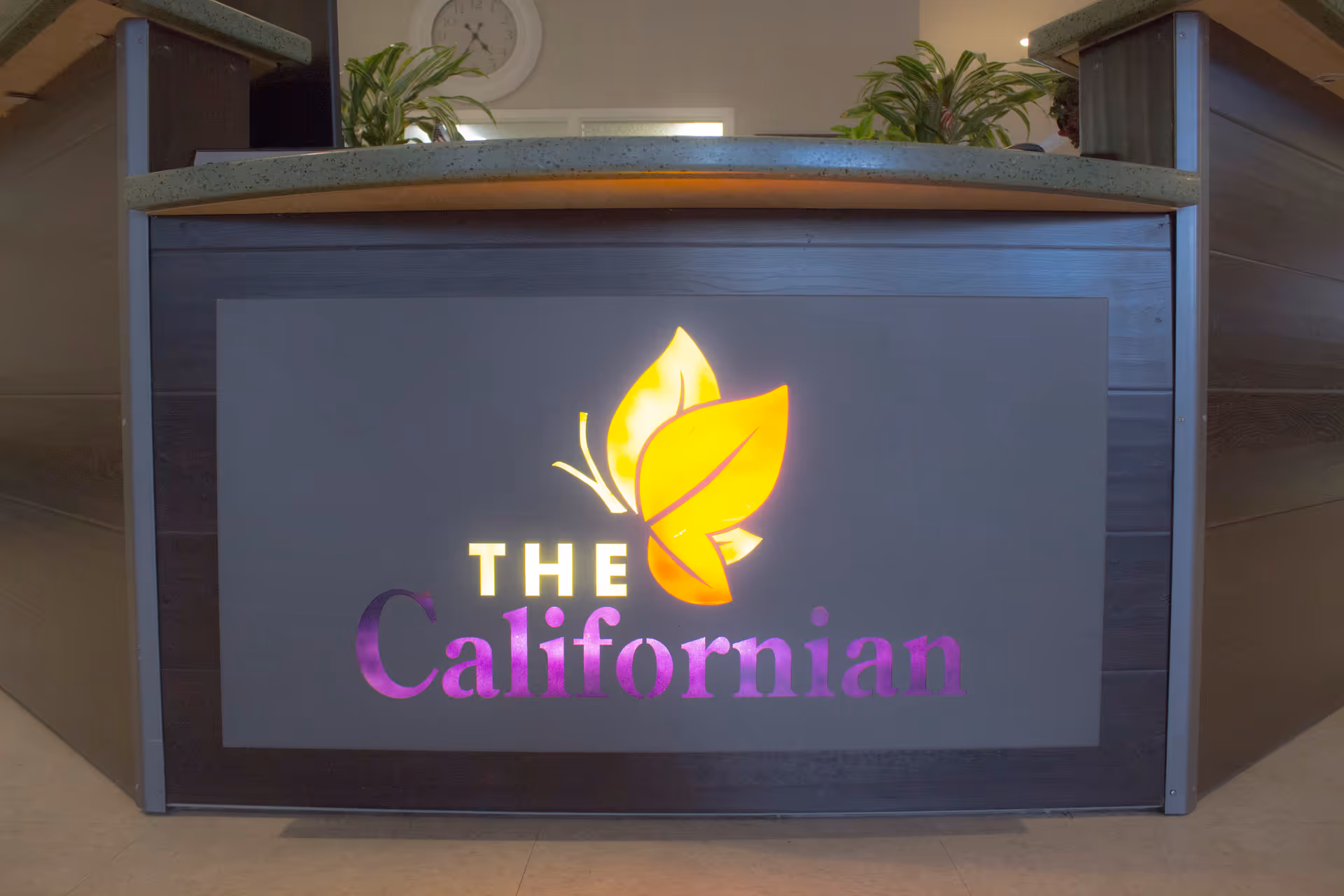 Reception desk with a lit sign displaying the logo and name 'The Californian' featuring a stylized leaf design, with plants and a clock visible in the background.